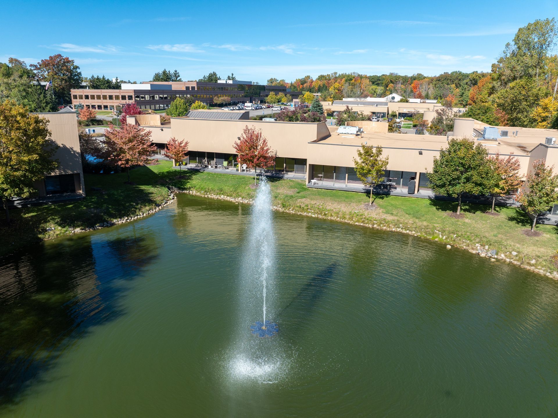 Pond with fountain in front of buildings and fall foliage under blue sky.