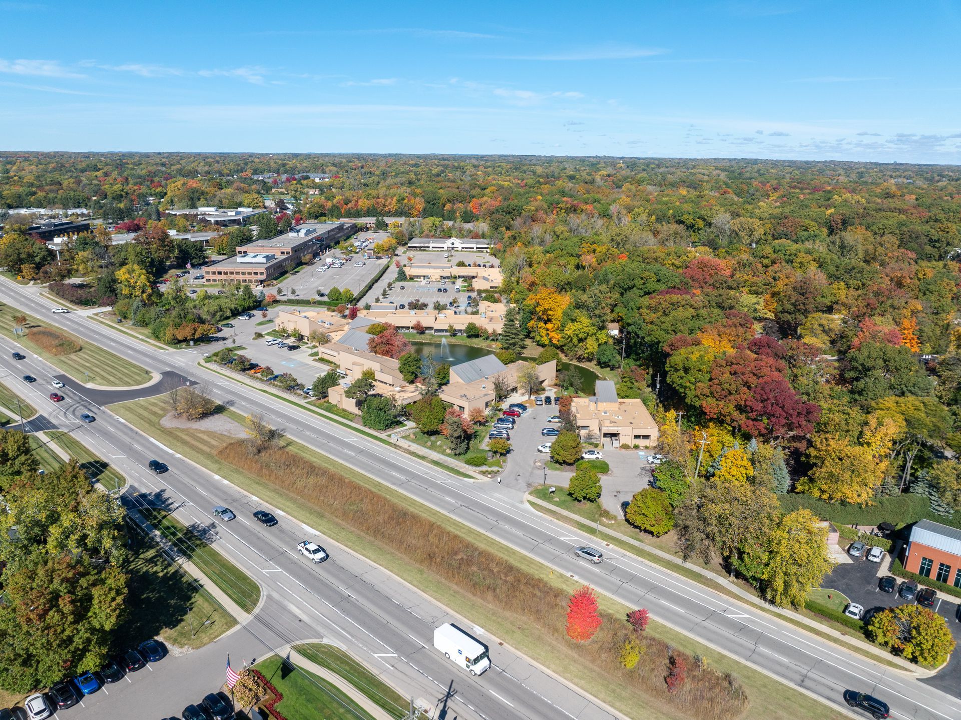Aerial view of buildings next to a highway, surrounded by colorful autumn trees and blue sky.