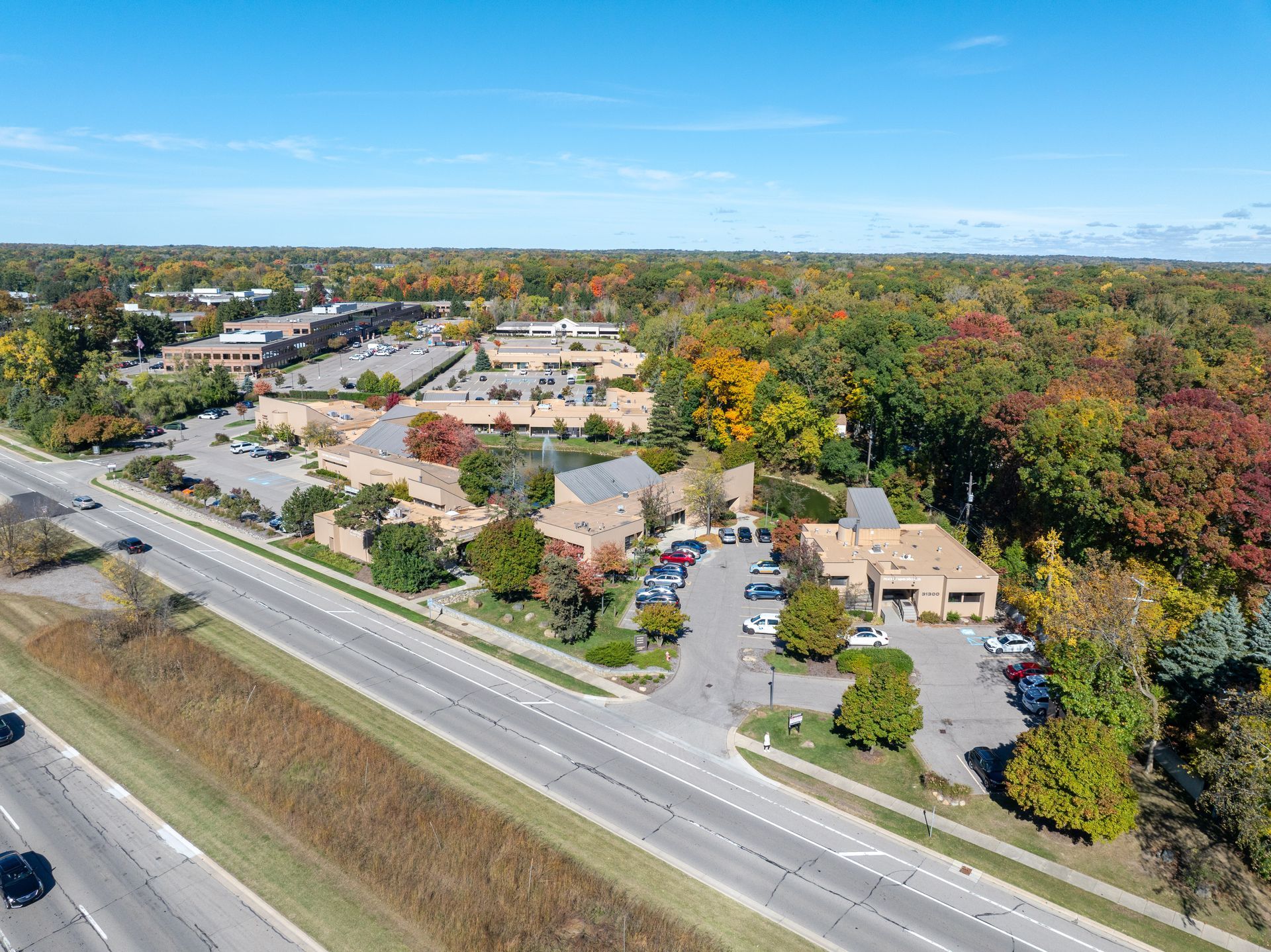 Aerial view of buildings, parking lot, and highway surrounded by colorful autumn trees under a blue sky.