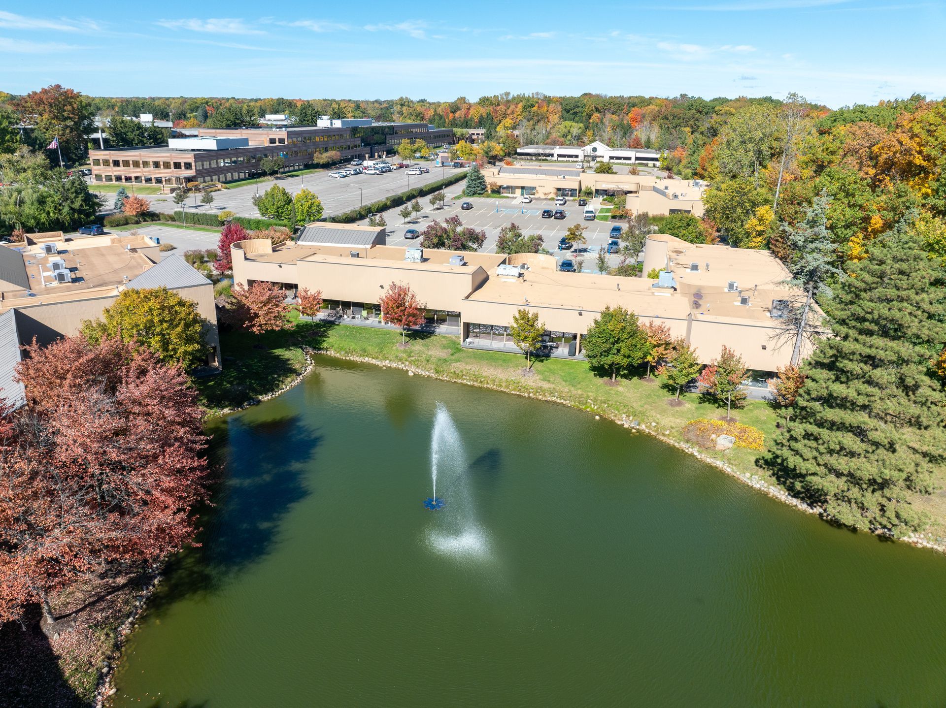 Aerial view of buildings near a pond with a fountain, surrounded by trees with fall foliage, under a blue sky.