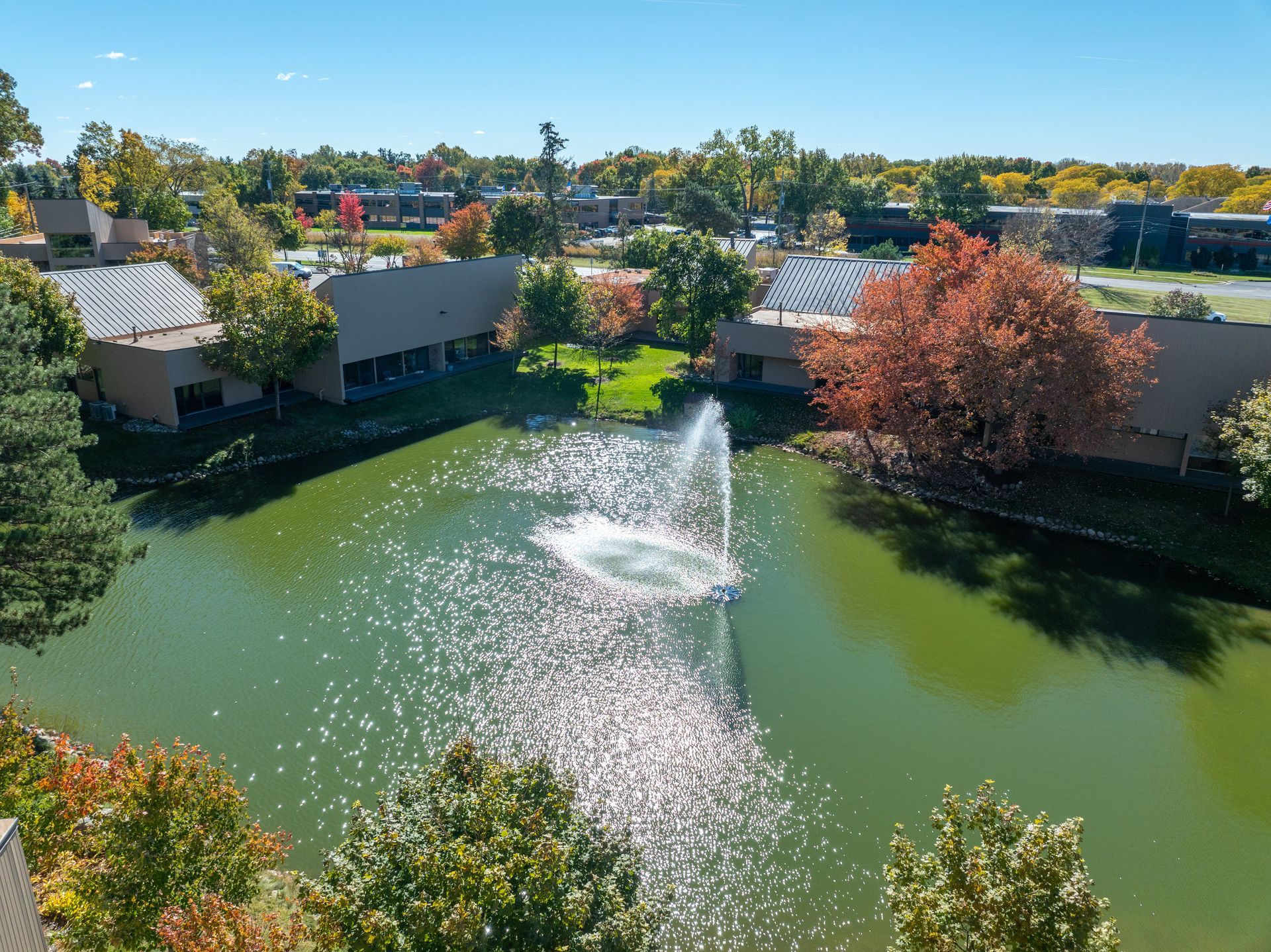 Pond with fountain spraying, surrounded by buildings and colorful fall trees under a clear blue sky.