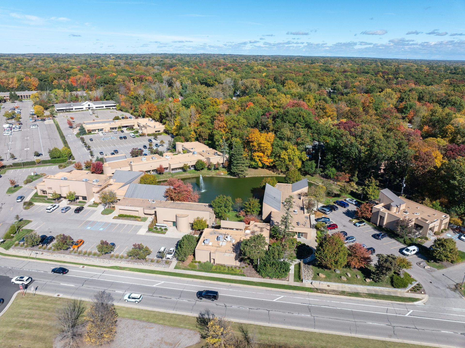 Aerial view of office buildings, pond, and colorful autumn trees beside a road and parking lots.
