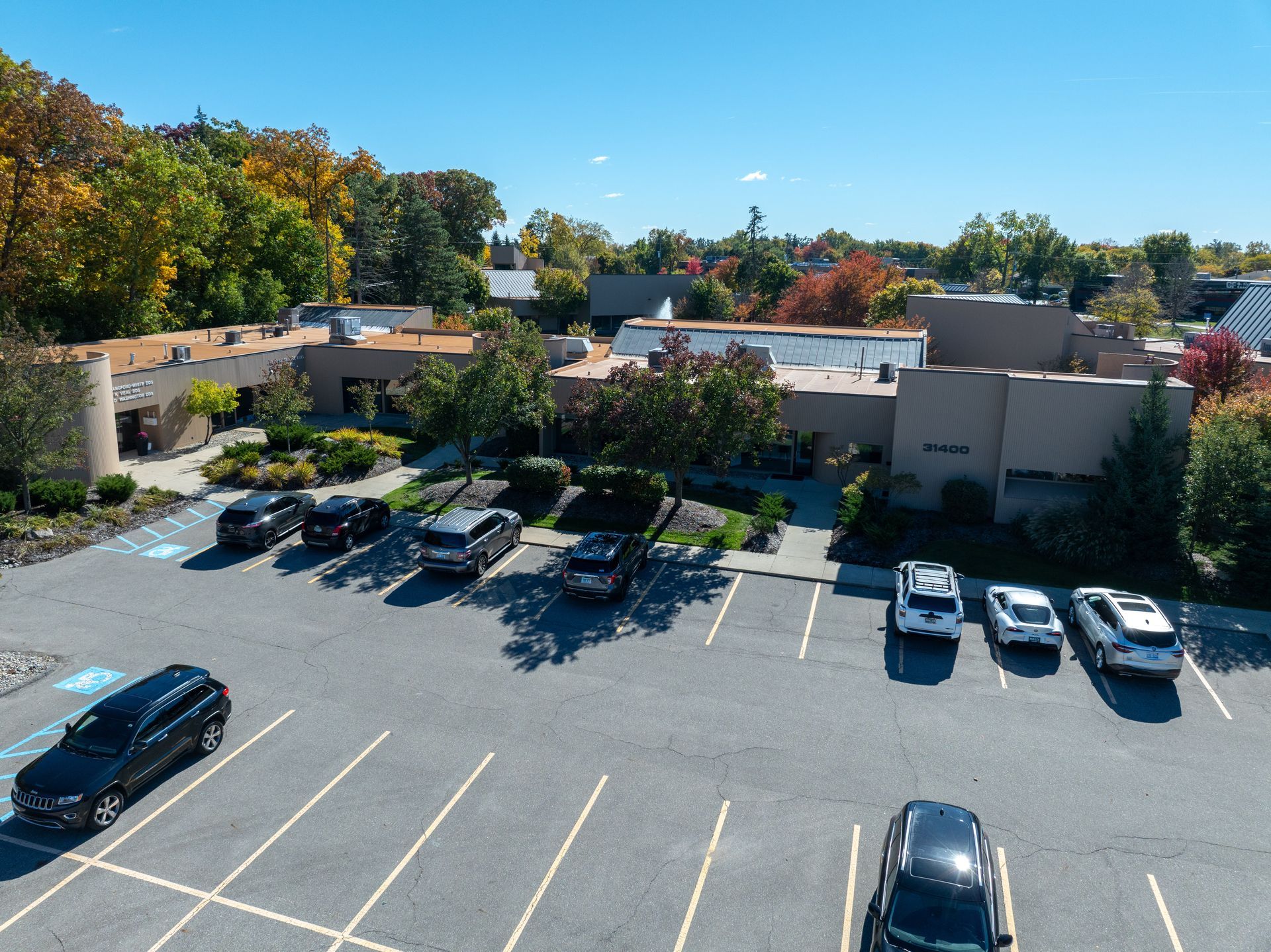 Parking lot with cars in front of a tan building with a flat roof, surrounded by fall foliage.