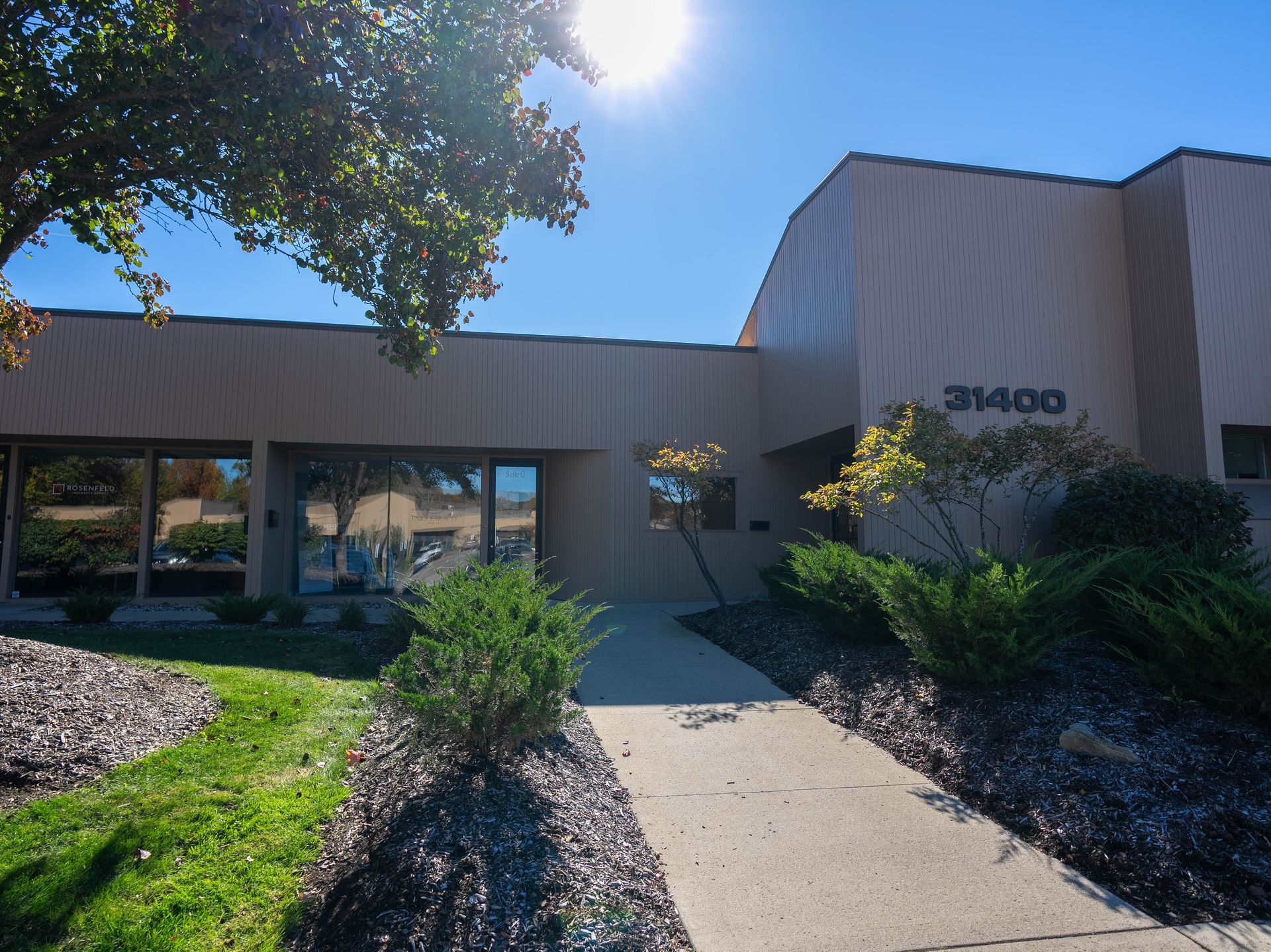 Tan building entrance with address 31400, walkway, bushes, blue sky, and sun.