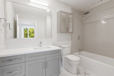 Modern bathroom with gray vanity, white tub, and toilet. Large mirror reflects a window