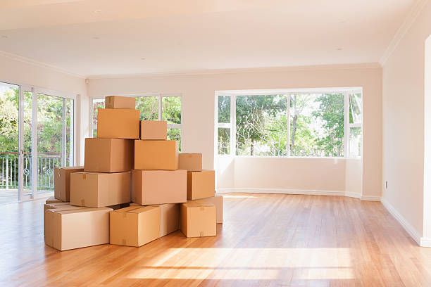 Boxes stacked in an empty room with a large window and wooden floor