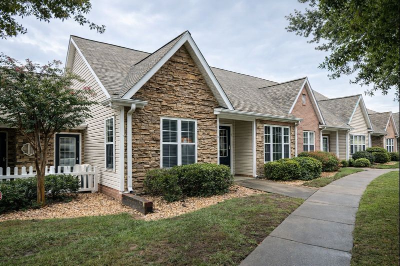 A row of single-story townhomes with light siding, stone facades, and gabled roofs, fronted by a sidewalk and landscaping.