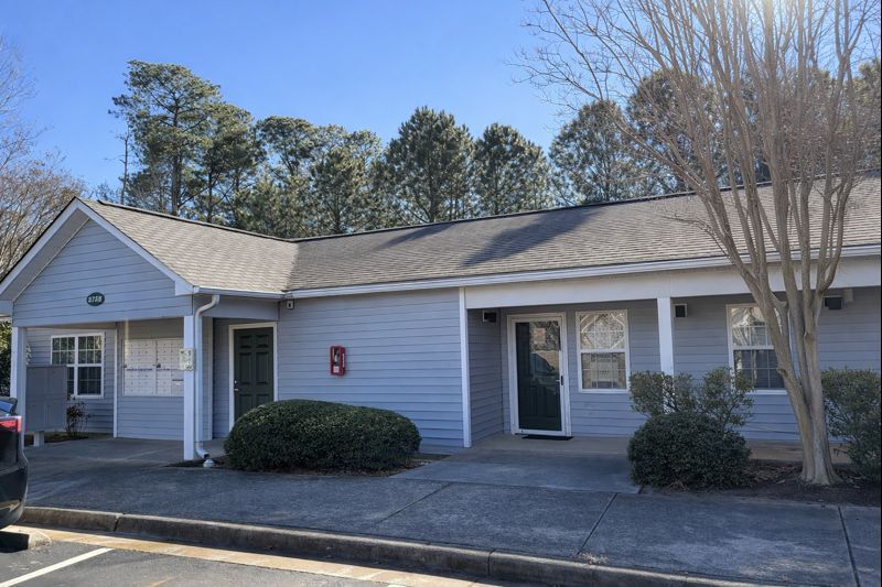 A light blue, one-story commercial building with a covered walkway, dark doors, and landscaping under a clear blue sky.