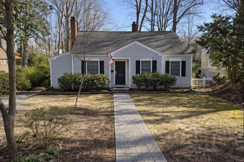 A single-story gray house with black shutters, a central front door, a brick chimney, and a stone walkway in a yard.