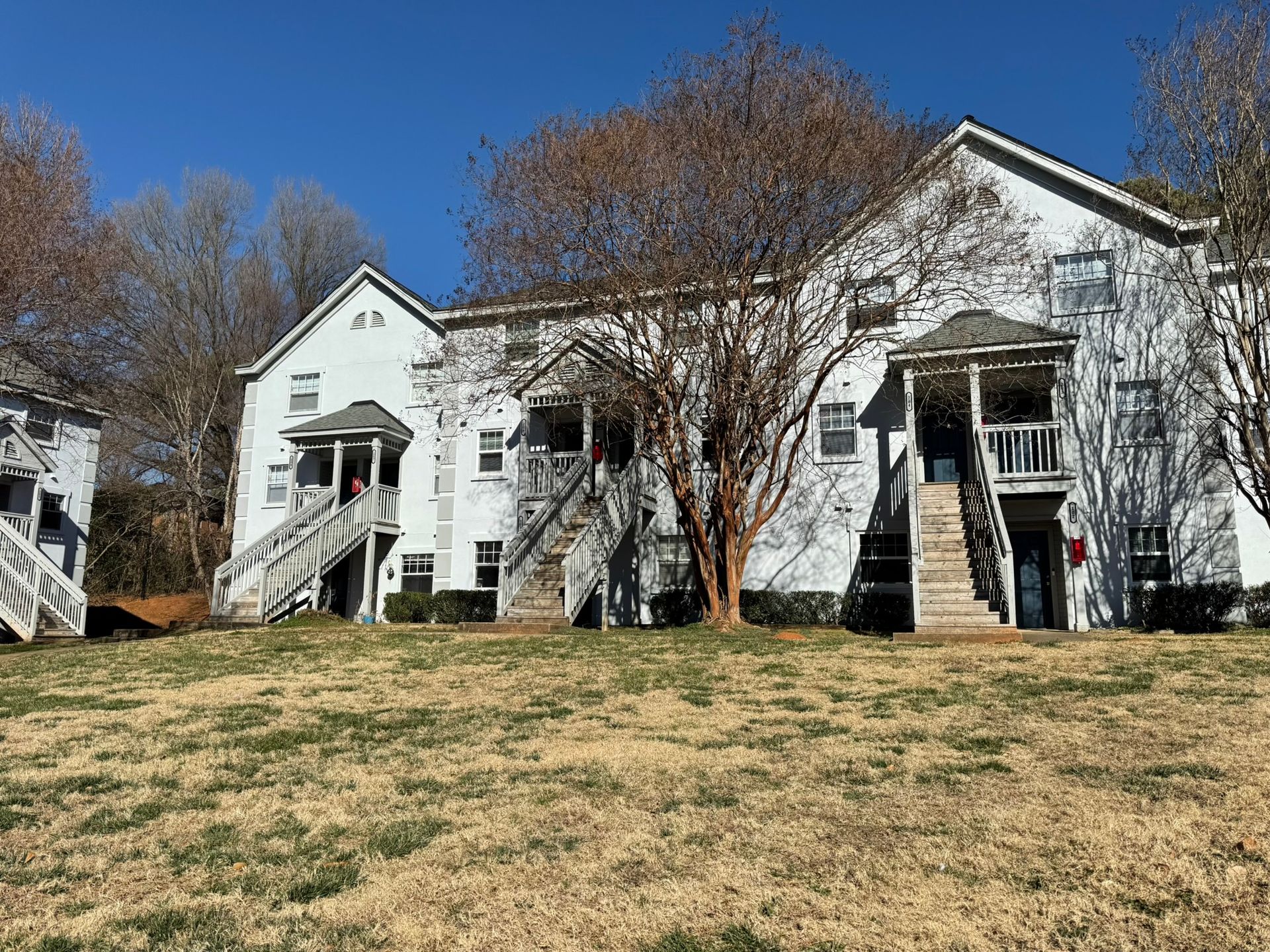 A white, two-story apartment complex with outdoor staircases, surrounded by bare trees under a clear blue sky.