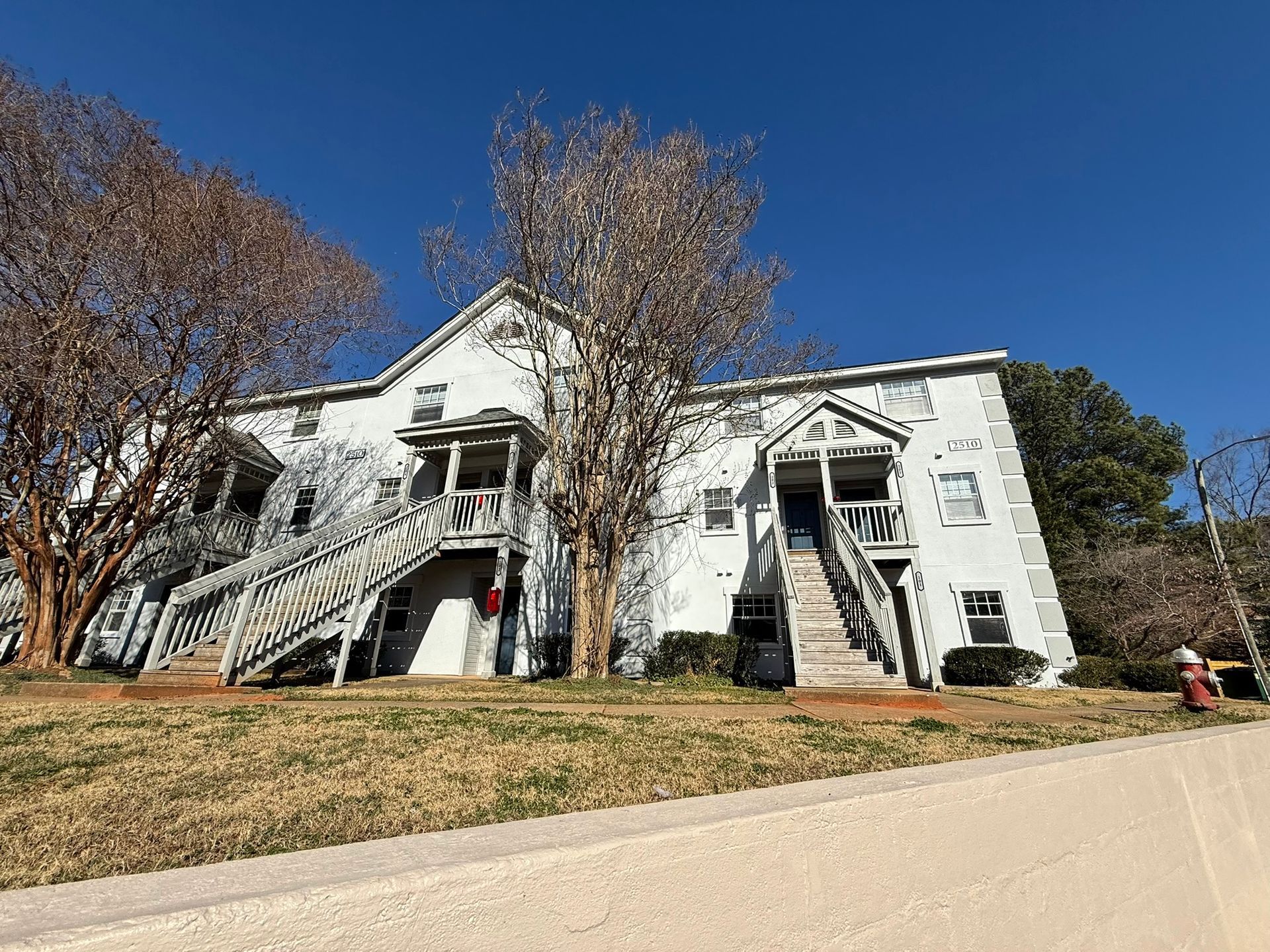 Two-story white apartment building with outdoor wooden staircases and bare trees under a clear blue sky.