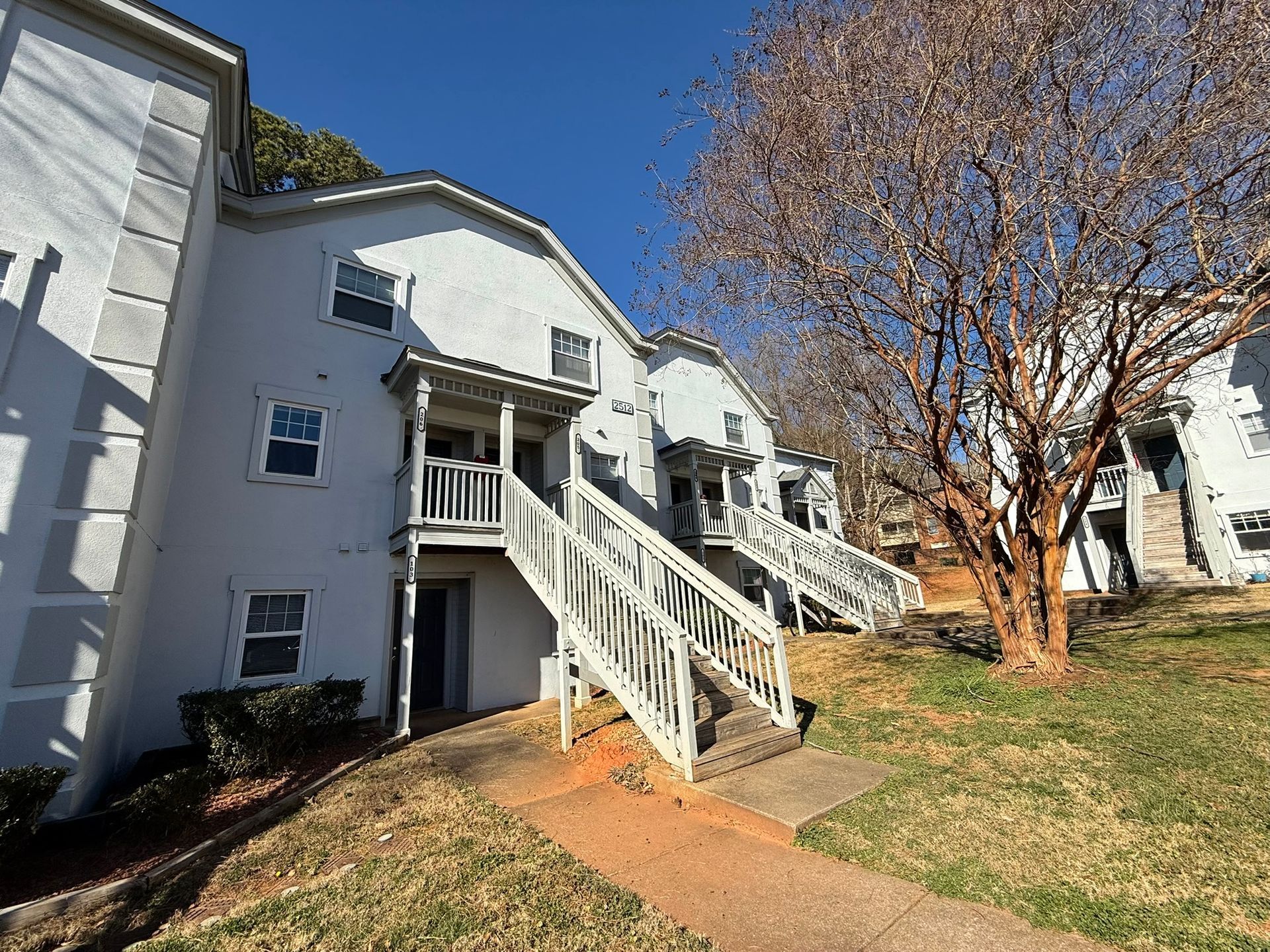 Two white, multi-story apartment buildings with wooden staircases and a bare tree on a sunny, grassy hillside.
