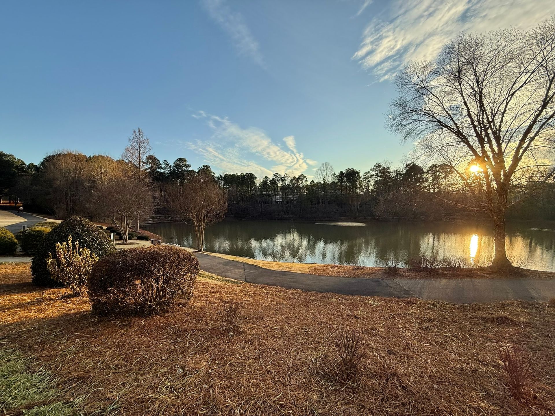A path winds along a calm pond at sunset, with golden light reflecting on the water and trees in an autumnal landscape.