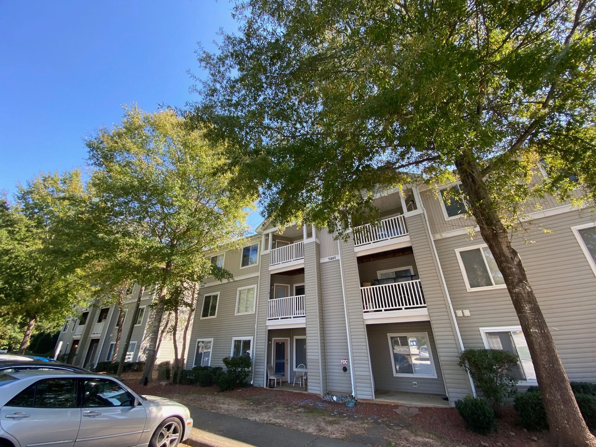 A beige three-story apartment building with balconies, surrounded by trees on a sunny day with a parked car in the front.