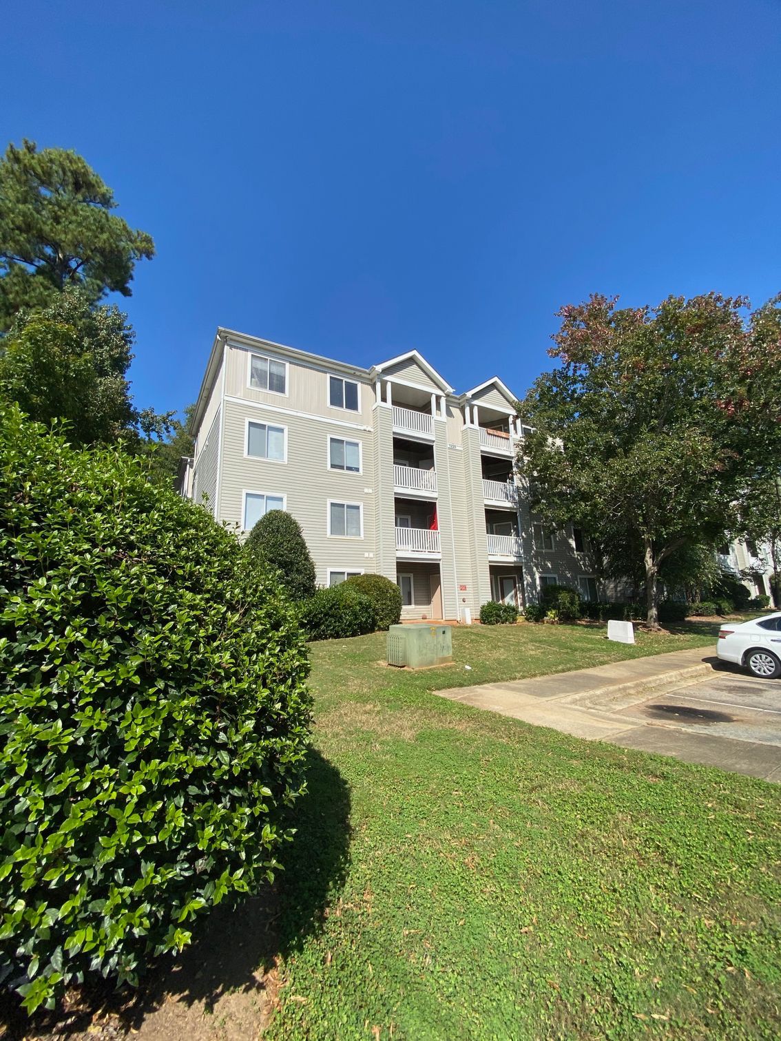 A four-story, light-colored apartment building stands under a clear blue sky, surrounded by trees and a green lawn.