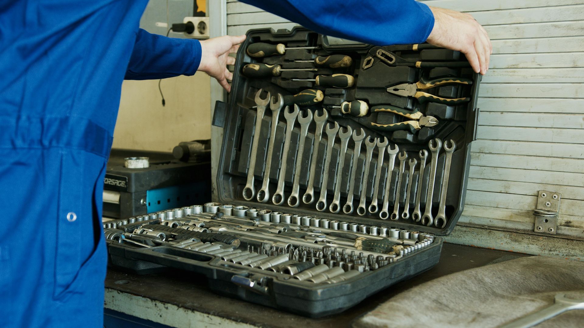 A man in a blue jumpsuit is opening a suitcase filled with tools.
