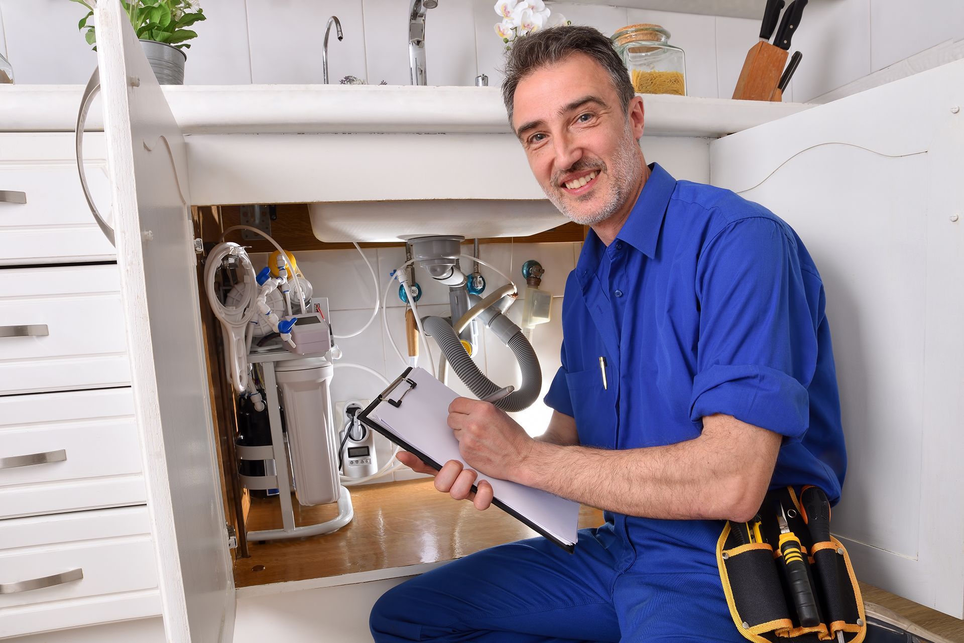 A plumber is kneeling under a sink in a kitchen holding a clipboard