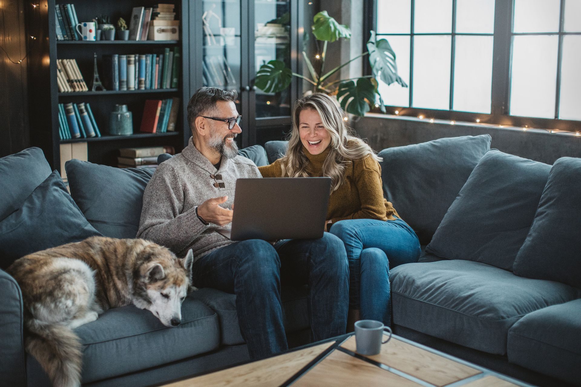 Couple laughing while looking at a laptop, sitting on a couch with a dog in a cozy living room.