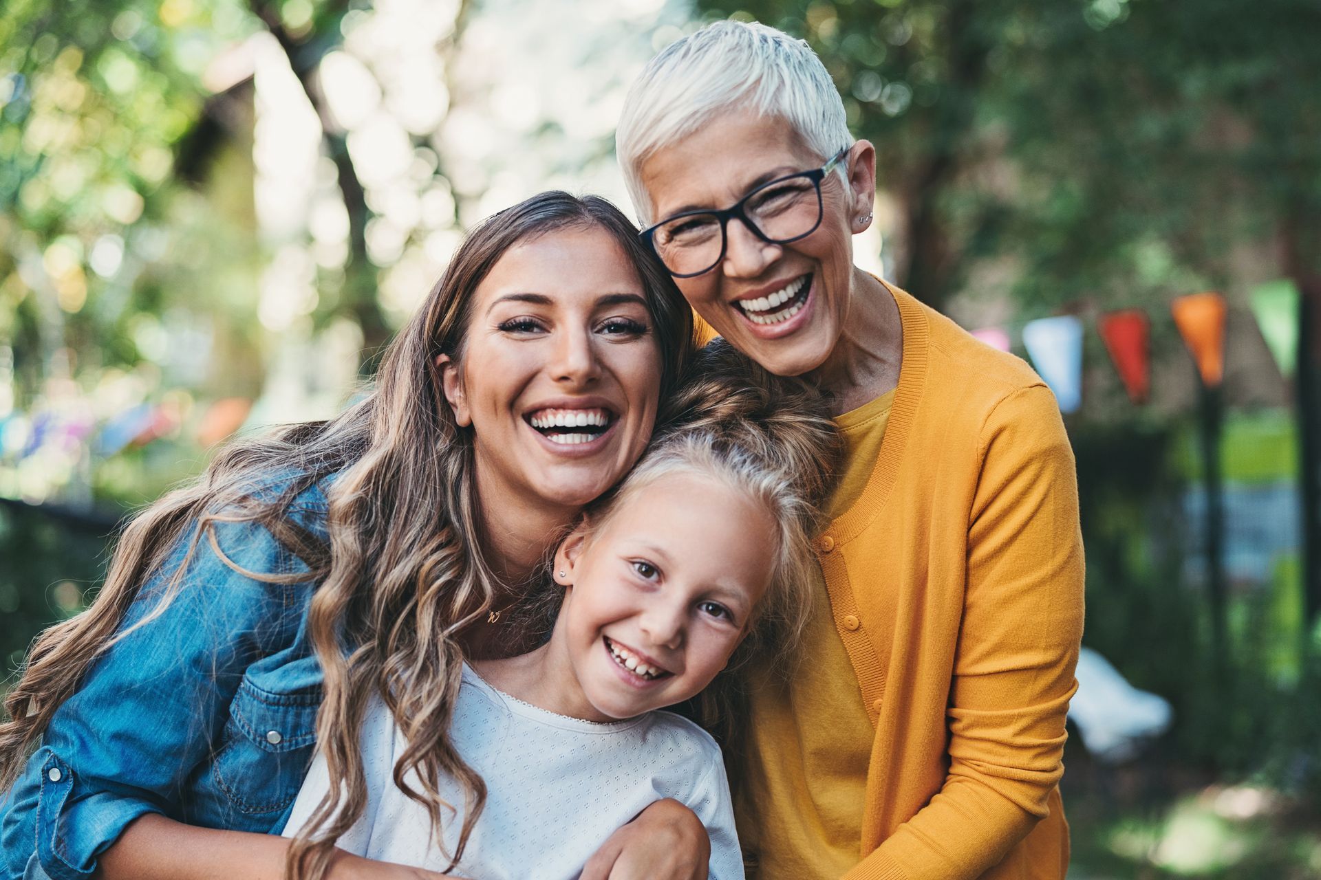 Three smiling women, embracing outdoors: a child, an adult, and an older woman in glasses.