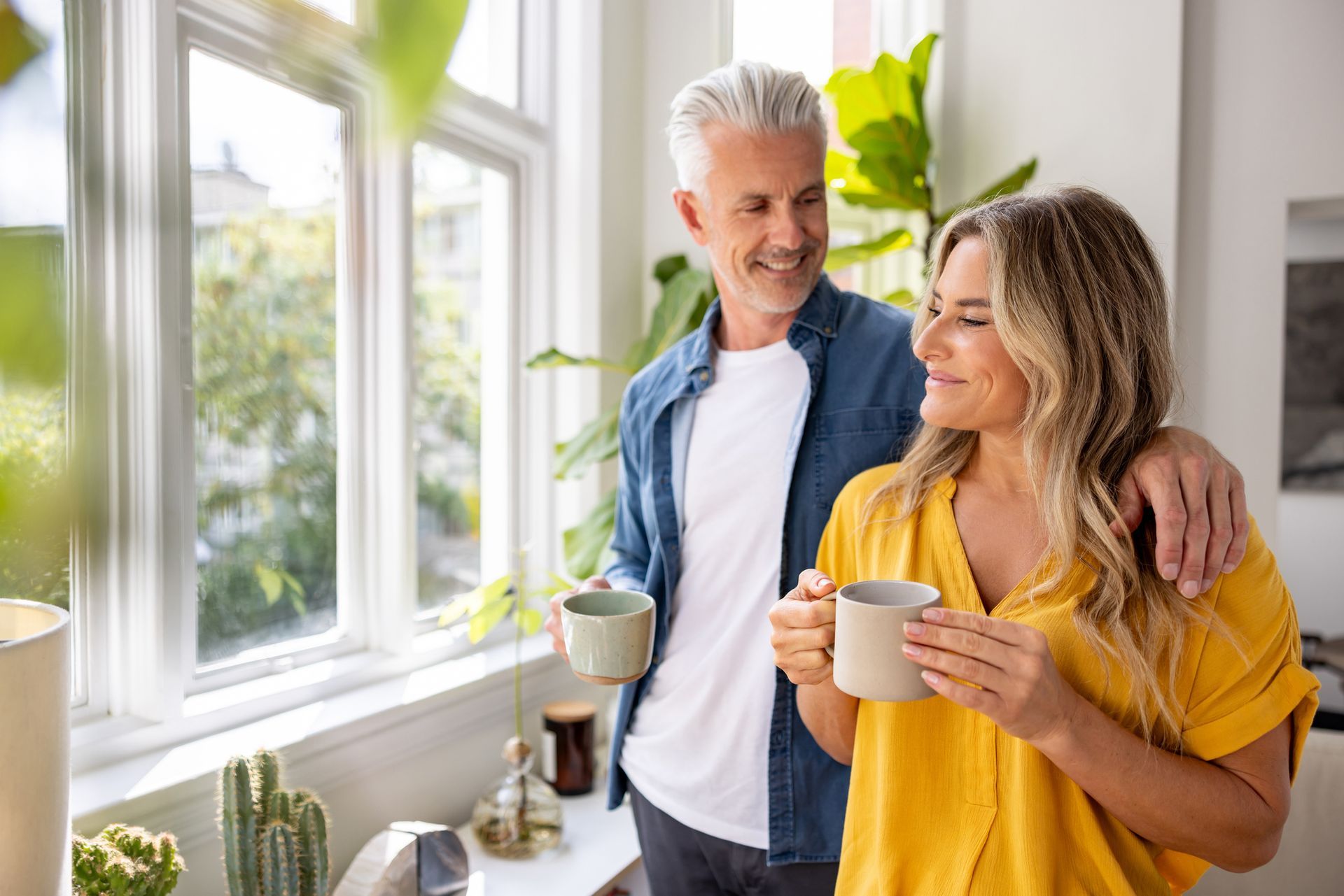 Couple with mugs of coffee, smiling by a window, arms around each other.