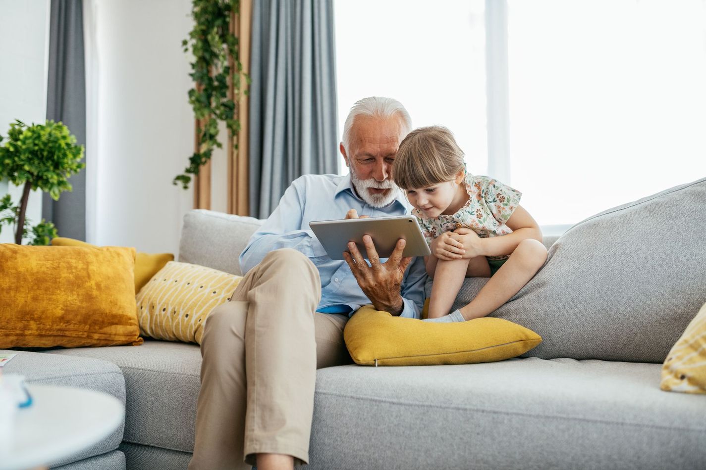 Grandfather and child looking at a tablet on a couch in a living room; yellow pillows.