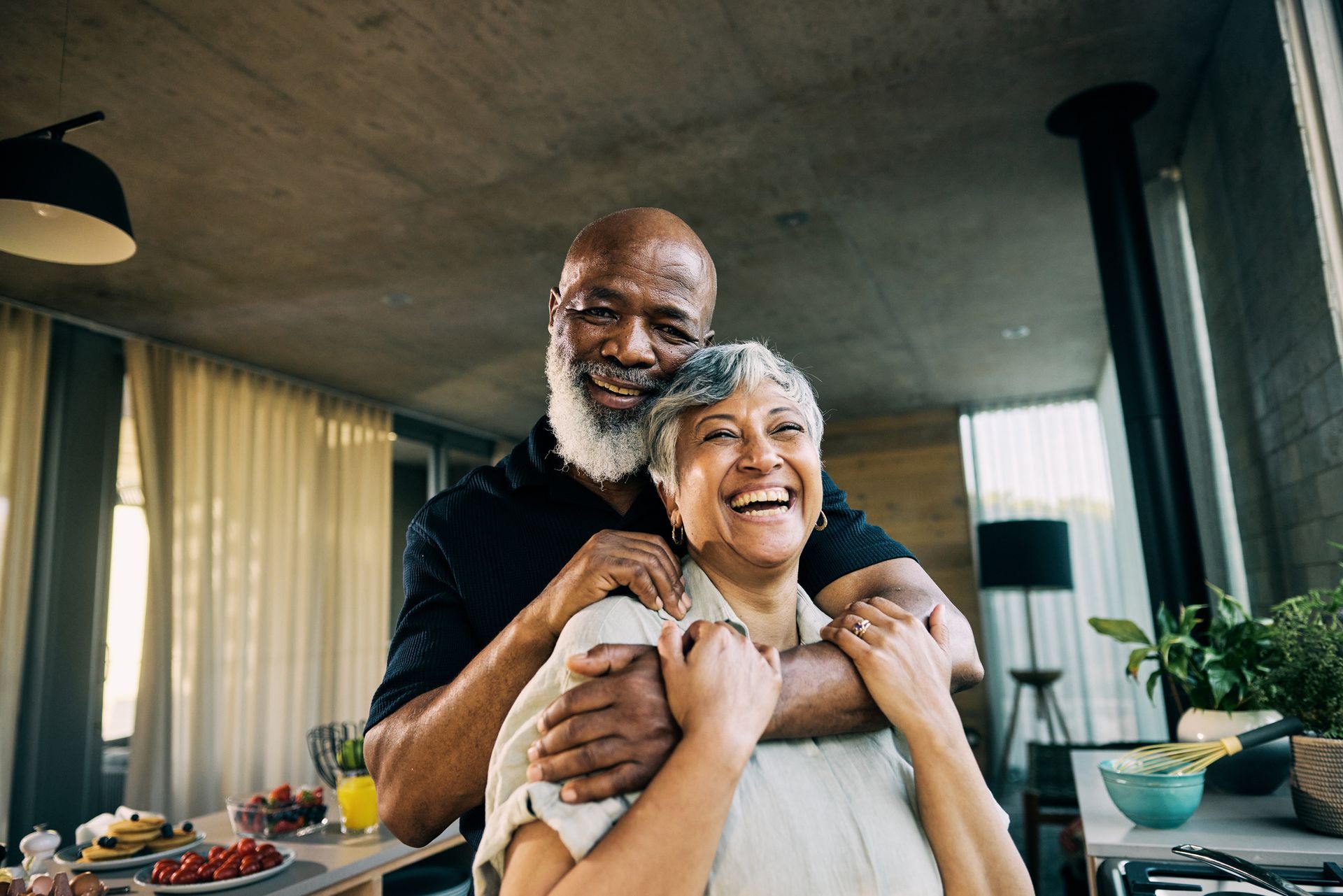 Smiling couple embracing indoors; man with beard, woman laughing, kitchen background.