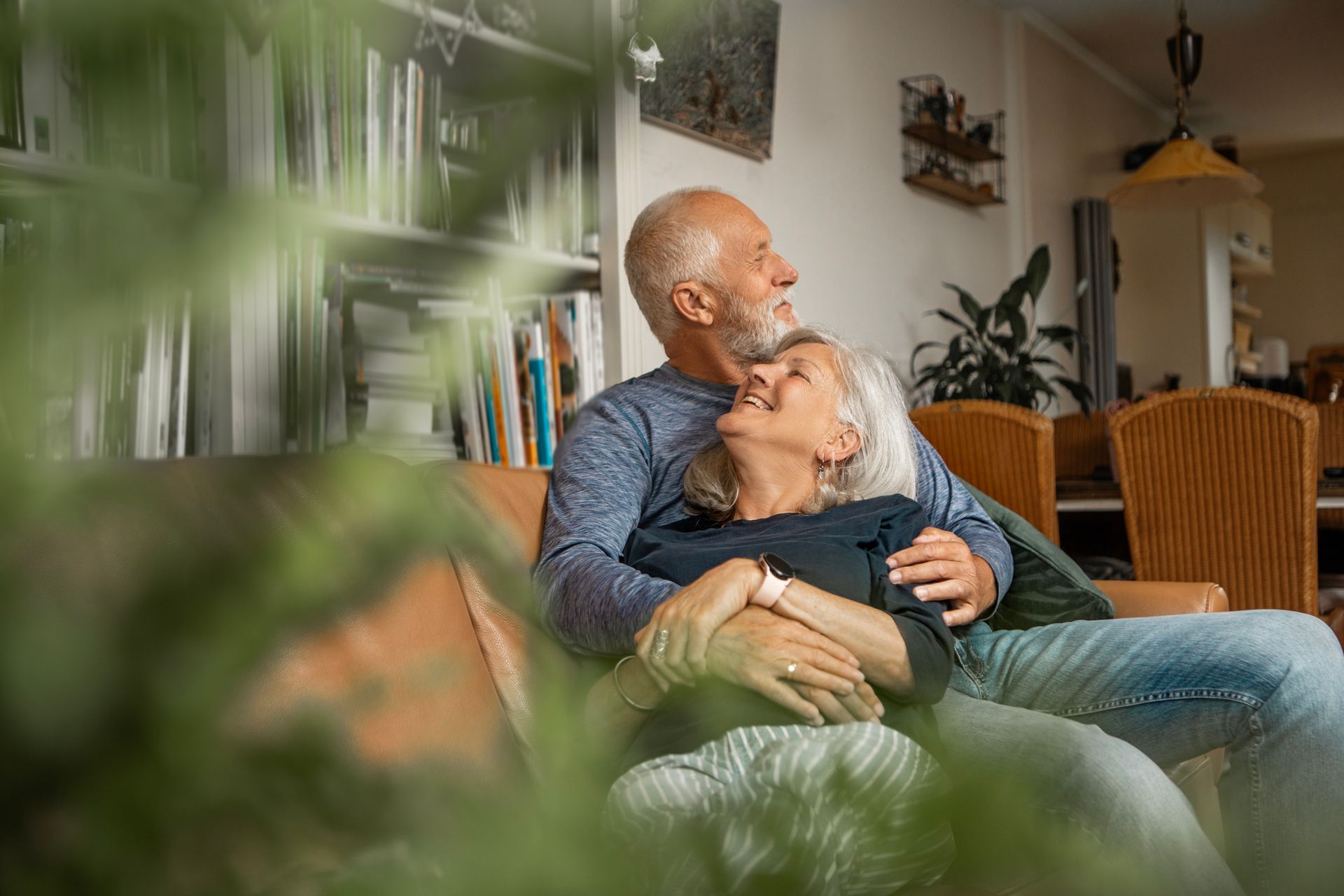 Older couple cuddling on a couch, looking upward and smiling in a well-lit living room.