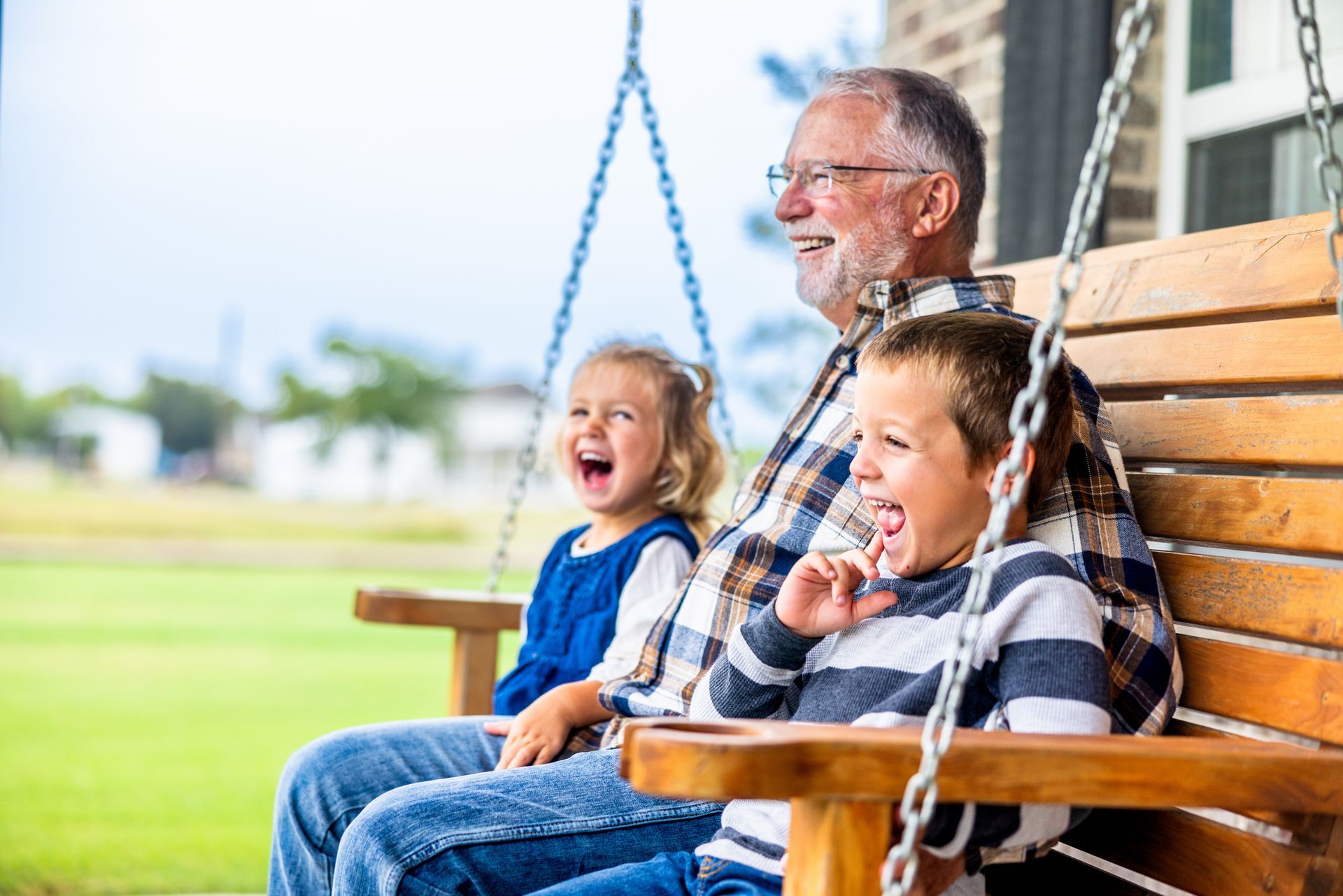 Grandfather and two grandchildren laughing on a wooden porch swing; blurred background of grass and sky.
