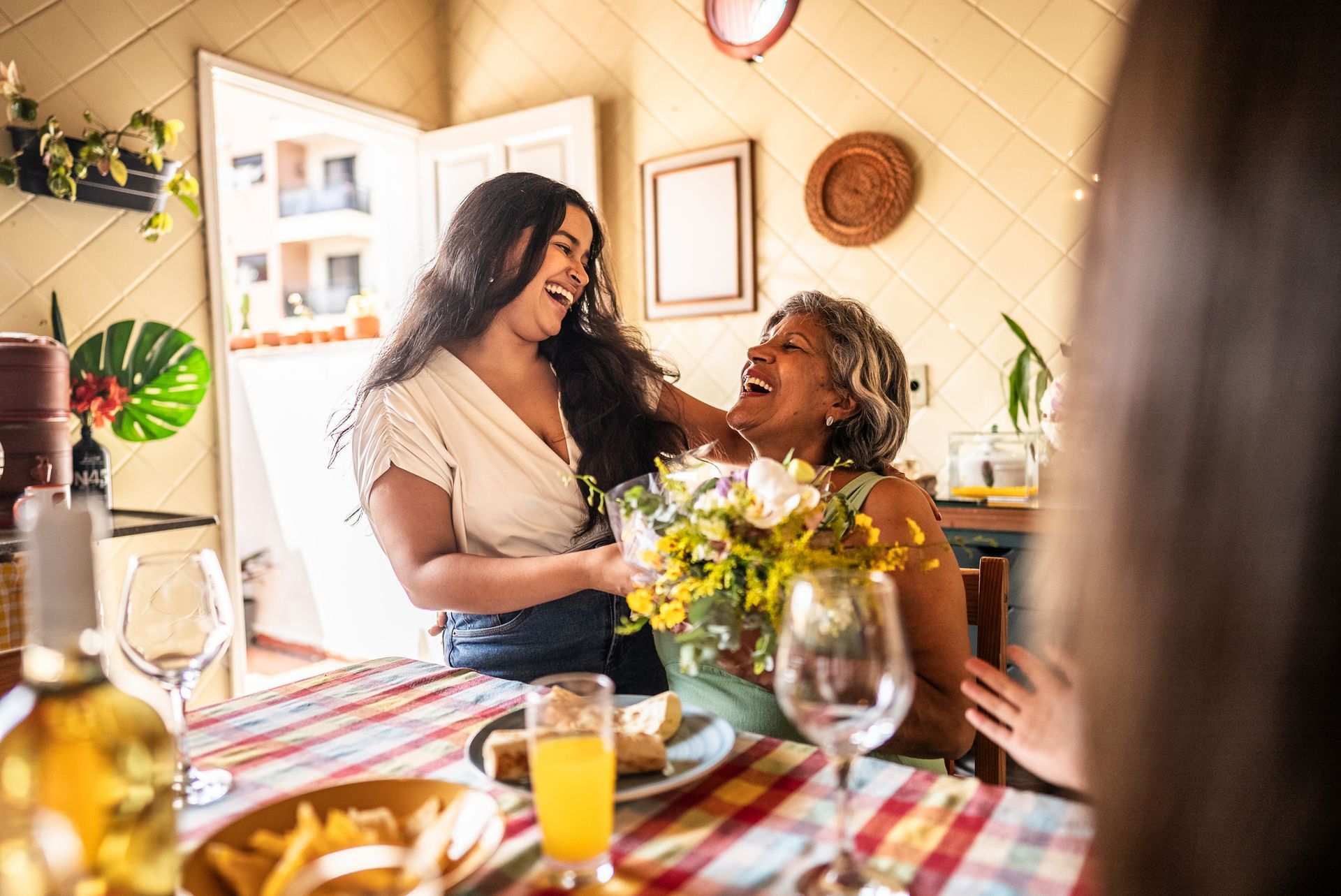 Woman laughing with older woman holding flowers at a table set for a meal.