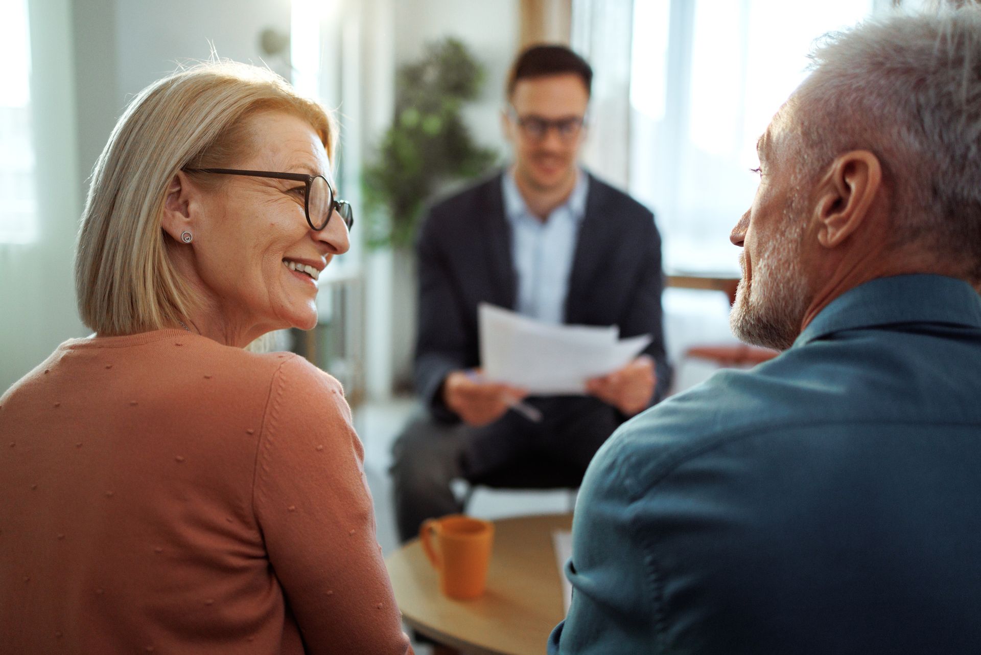 Elderly couple meeting with a financial advisor, smiling, discussing documents in a bright living room.