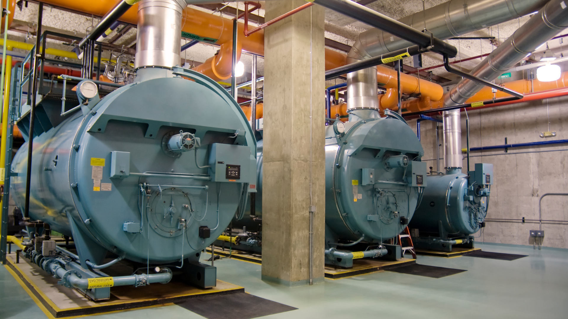 Three boilers are lined up in a room with pipes hanging from the ceiling.