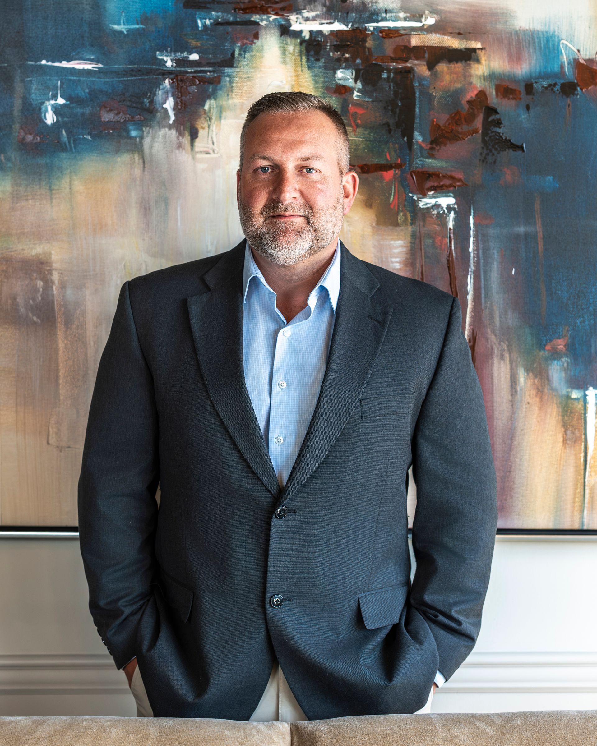 Luke Patton in a suit and blue shirt is standing in front of a painting.