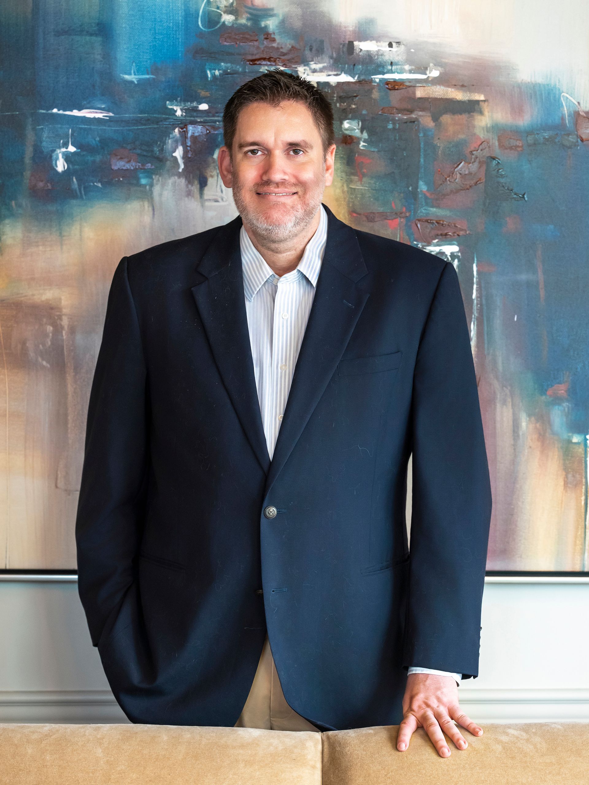 A man in a suit and blue shirt is smiling in front of a painting.