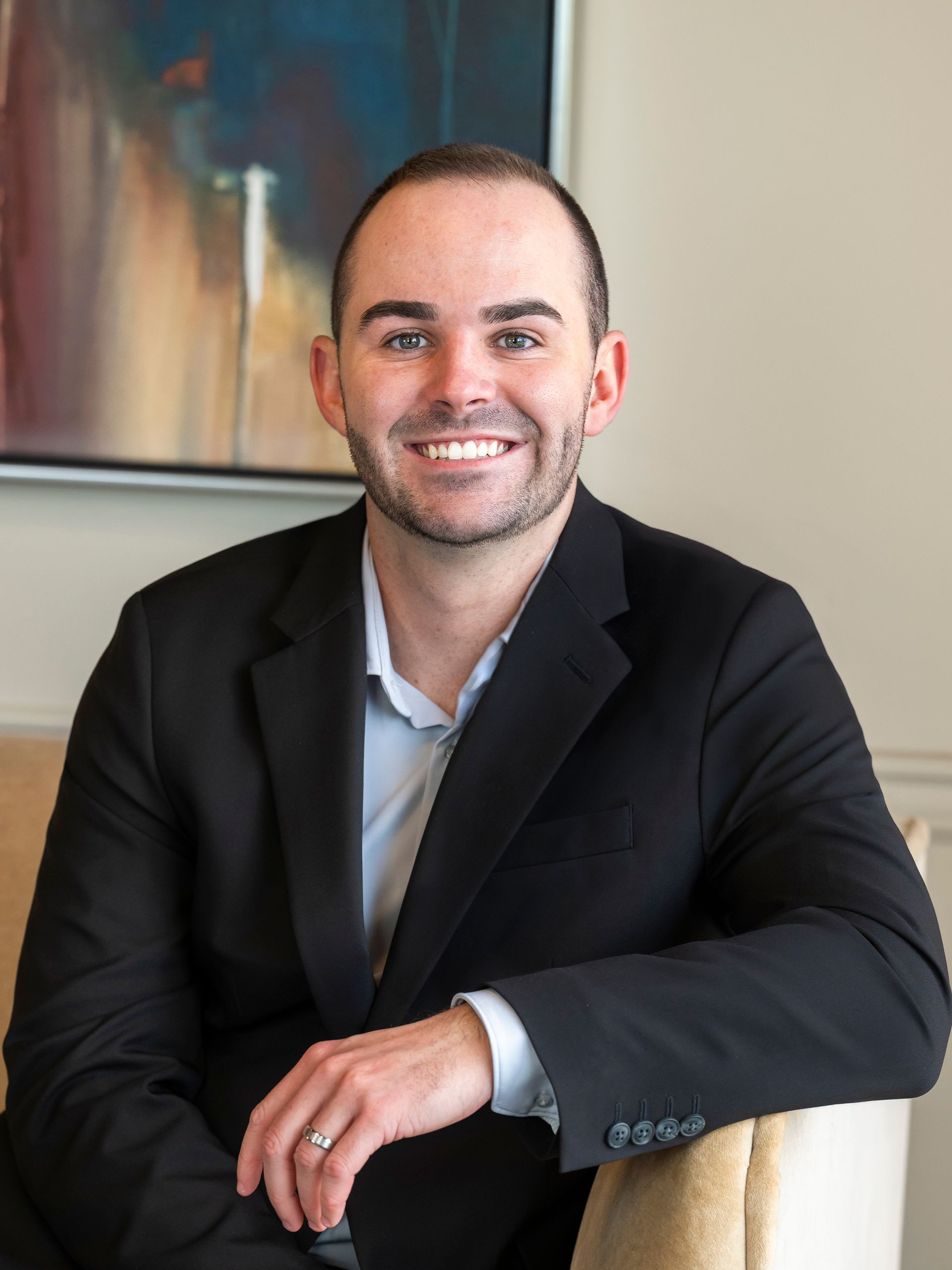 A man in a suit and blue shirt is smiling in front of a painting.