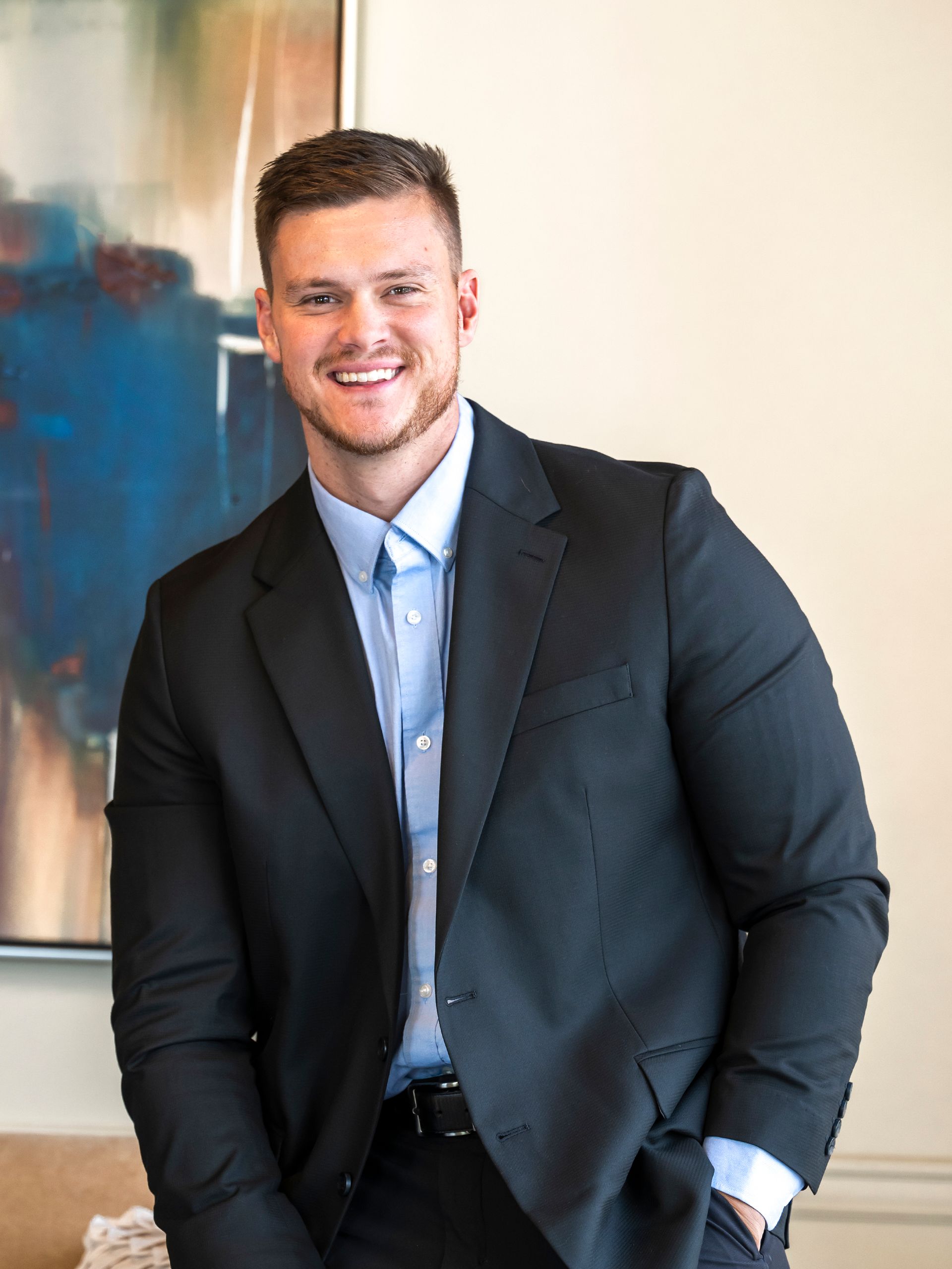 A man in a suit and blue shirt is smiling in front of a painting.