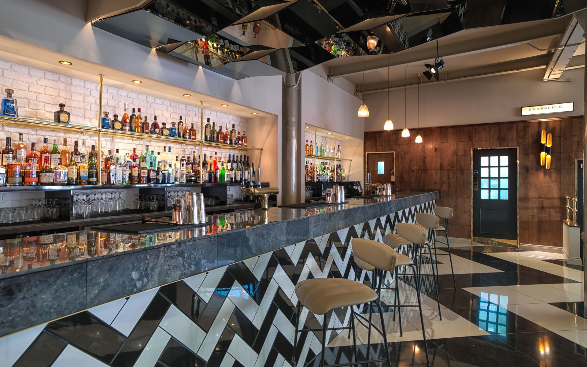 Bar interior with a marble countertop and chevron floor. Shelves hold bottles. Bar stools sit at the counter.