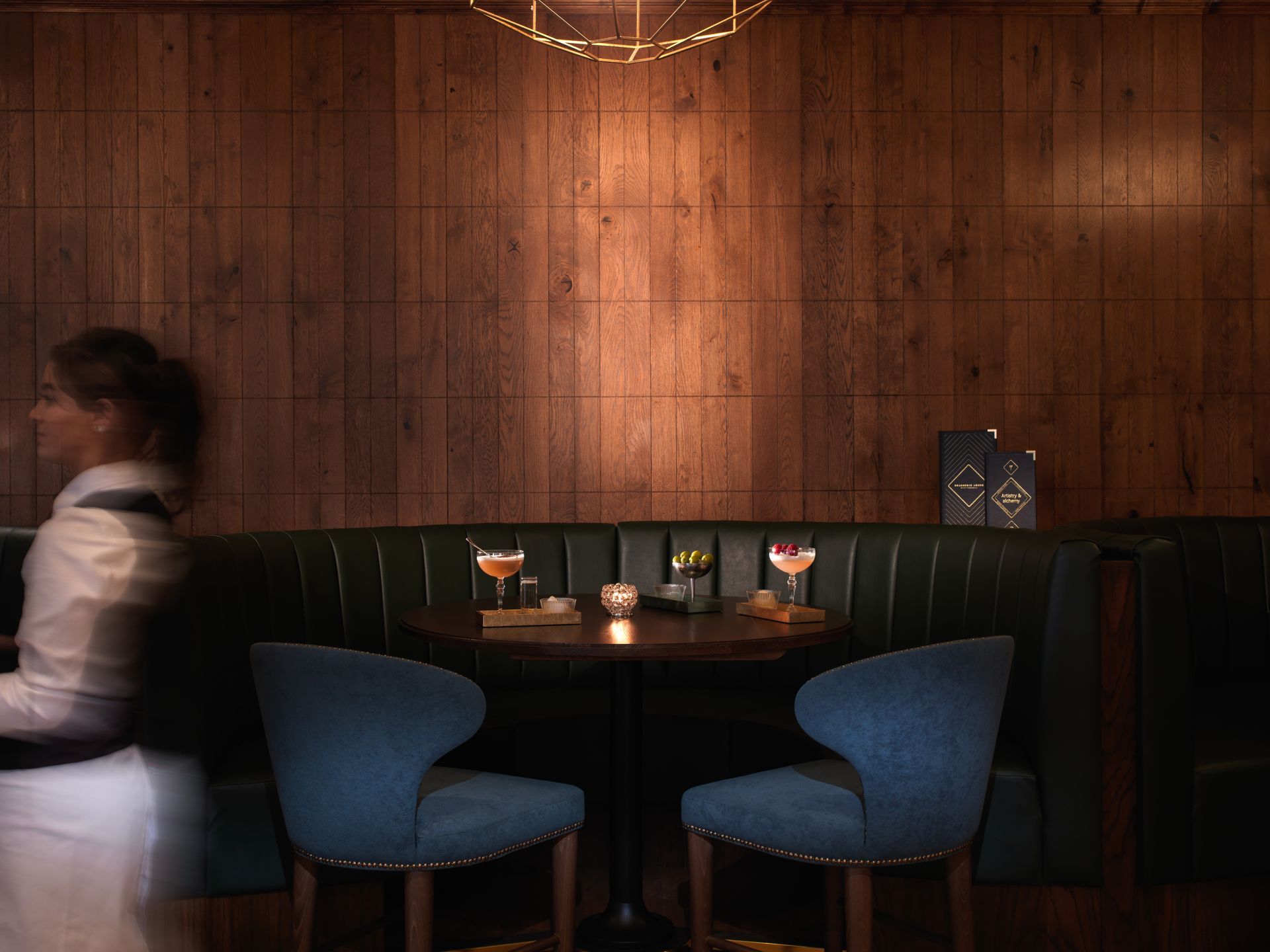 Dark wood-paneled booth in a bar with blue chairs, two cocktails, and a blurred waiter.