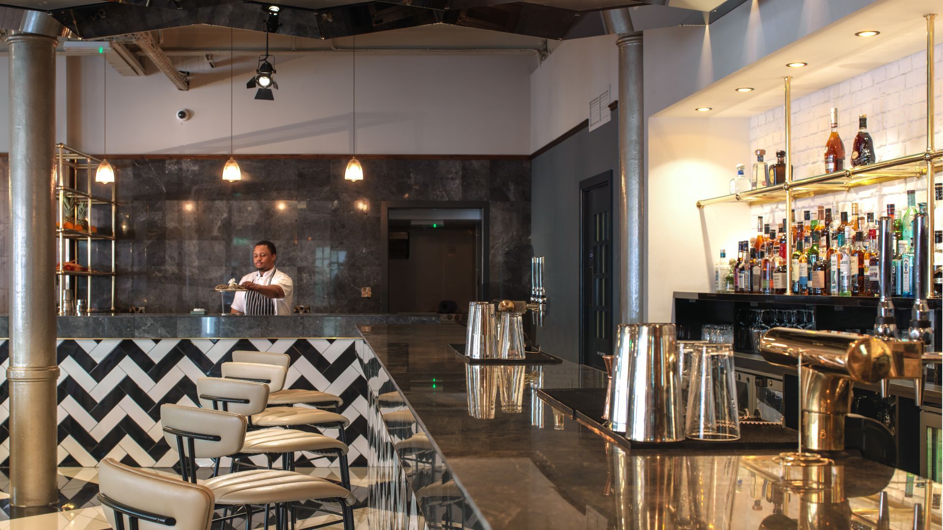 A bartender making a drink behind a modern bar with black and white chevron tile, gold accents, and bottles.