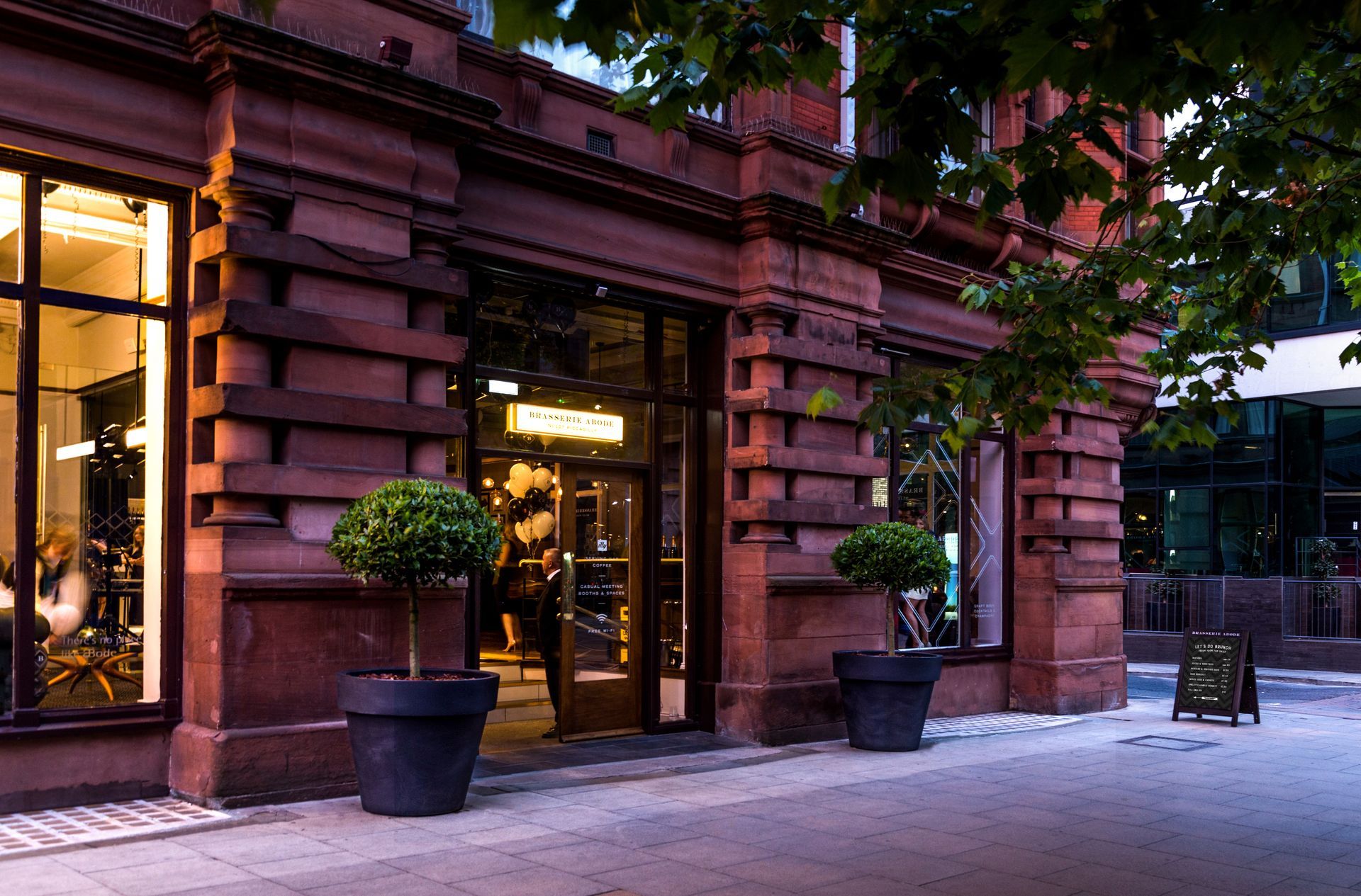 Restaurant entrance with dark glass door, brown stone facade, potted trees, and a sidewalk.
