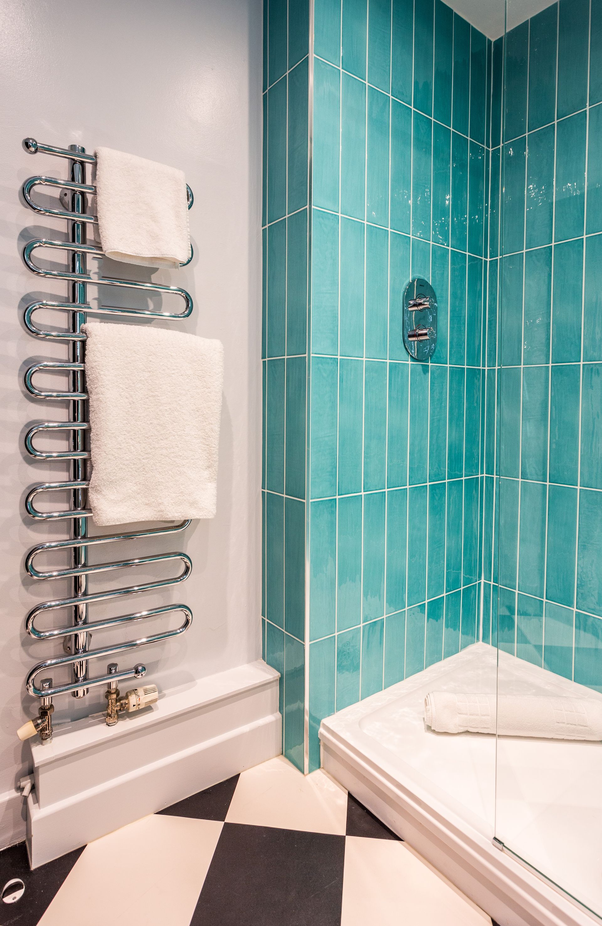 Bathroom with teal tiled shower, chrome towel rack with white towels, and black and white checkered floor.