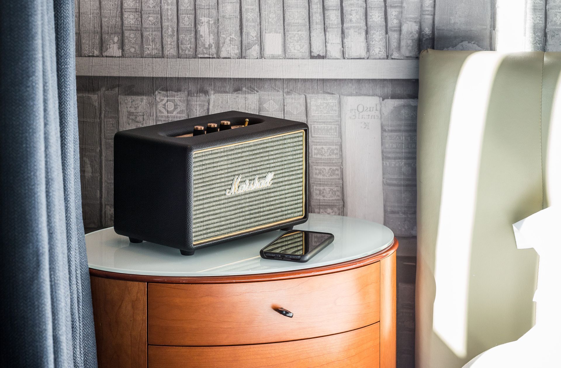 Black Marshall speaker on a nightstand with a phone. Wall background is books.