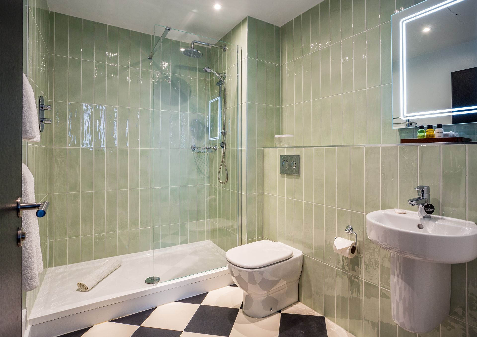 Bathroom with green tiled walls, glass shower, pedestal sink, toilet, and checkered floor.