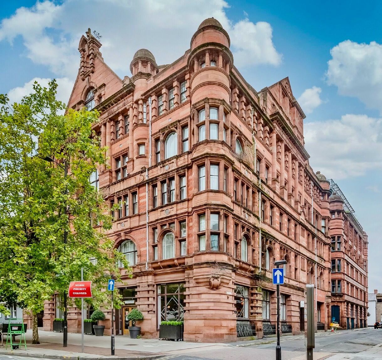 Red brick building with multiple floors, arched windows, and rounded corner turrets on a city street under a blue sky.