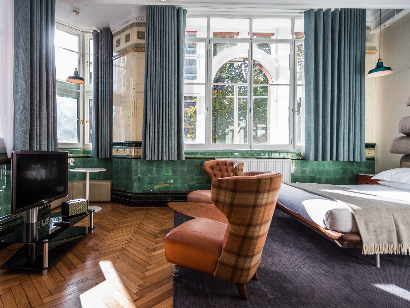 A stylish hotel room with a chair, chaise lounge, TV, and arched window with teal curtains.