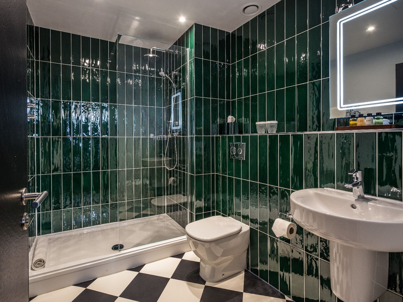 Green-tiled bathroom with black and white checkered floor, toilet, sink, shower, and mirror.