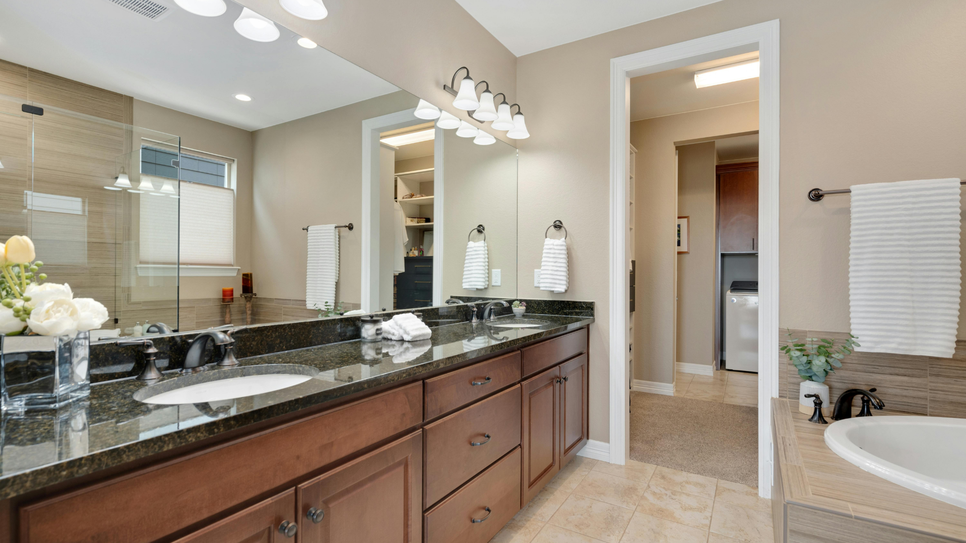Bathroom with double sink vanity, dark countertops, brown cabinets, neutral walls, and a view of a laundry room.