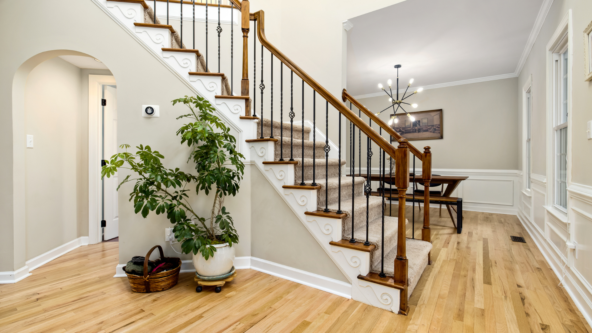 Wooden staircase with brown railings, leading up. Light-colored walls, wood floors, and a large houseplant.