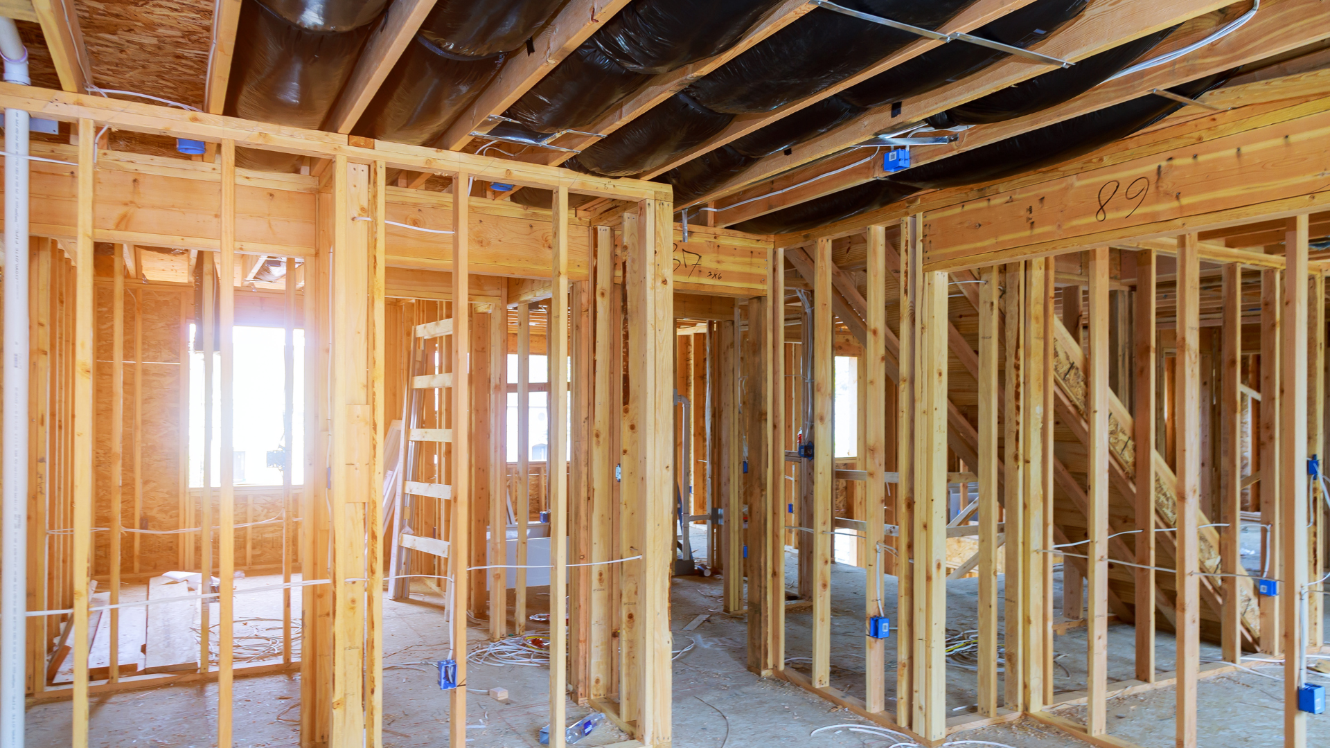 Interior framing of a house under construction; wooden studs, ceiling ductwork, window, and blue electrical boxes.