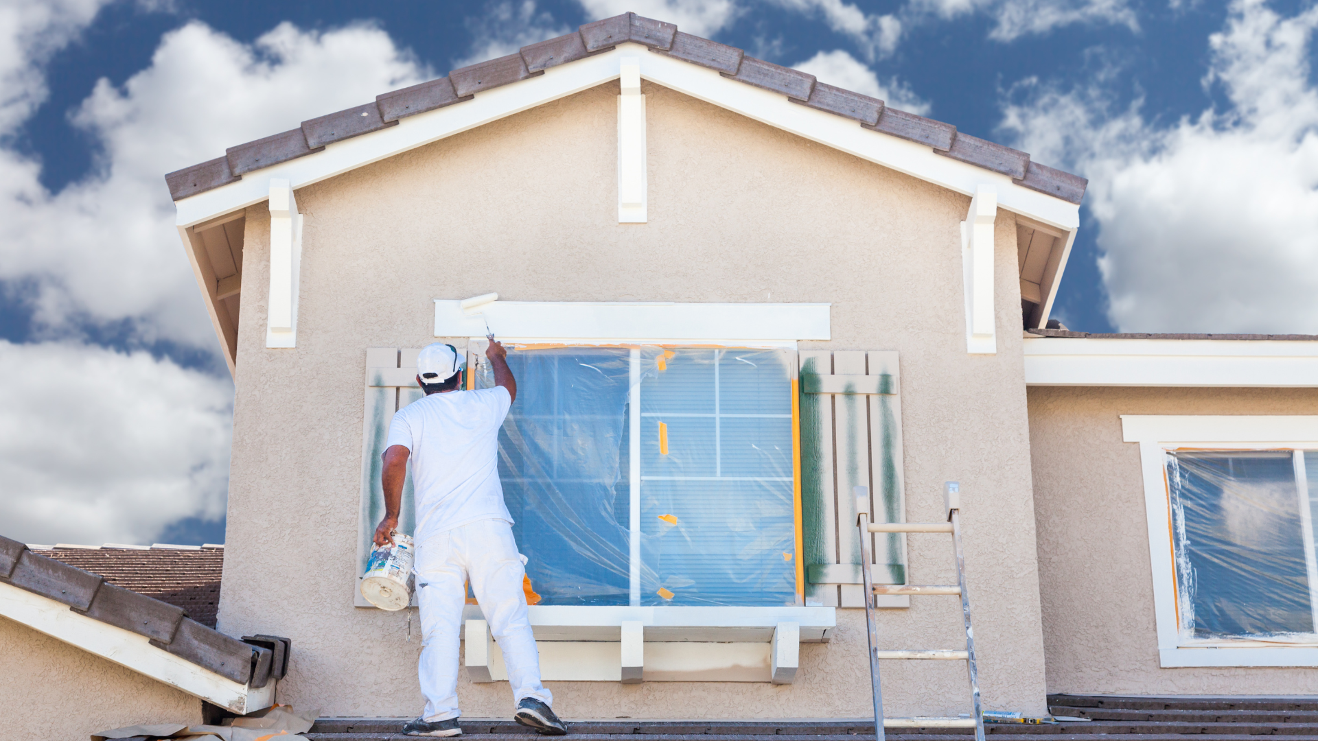 Painter in white overalls painting the exterior of a house, blue sky background.