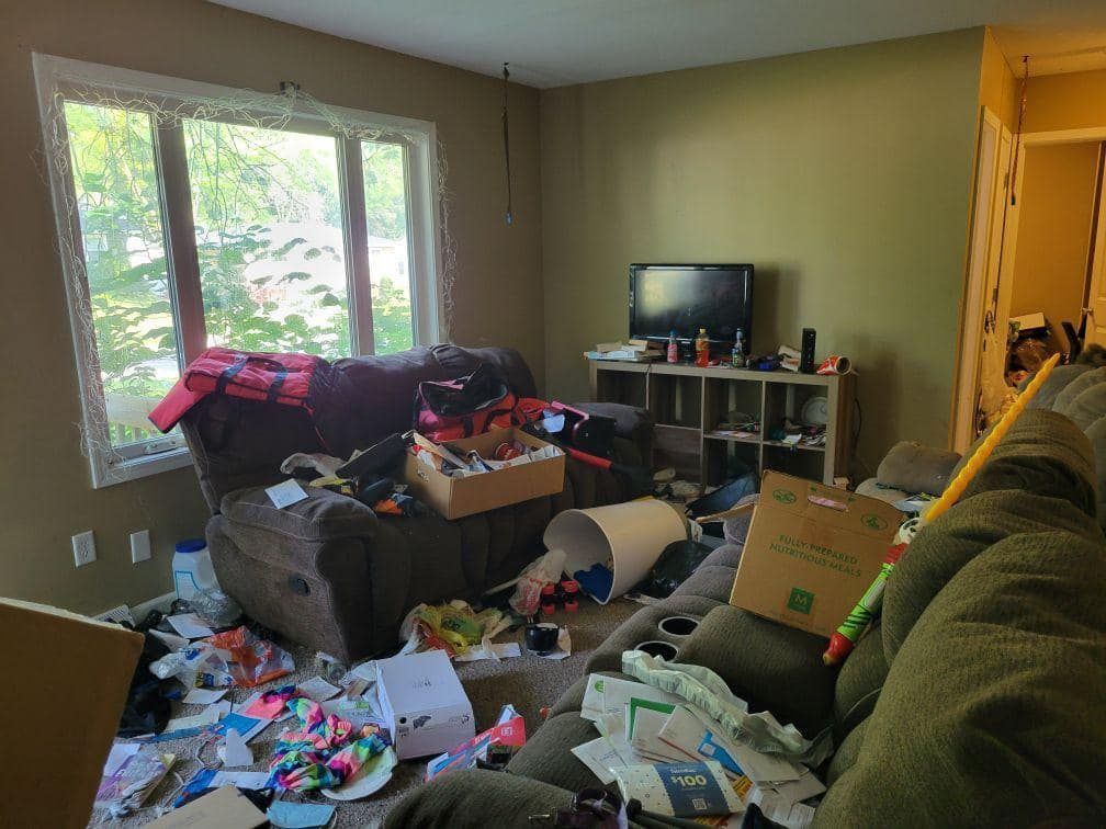 Cluttered living room with scattered papers, boxes, clothes, and miscellaneous junk
